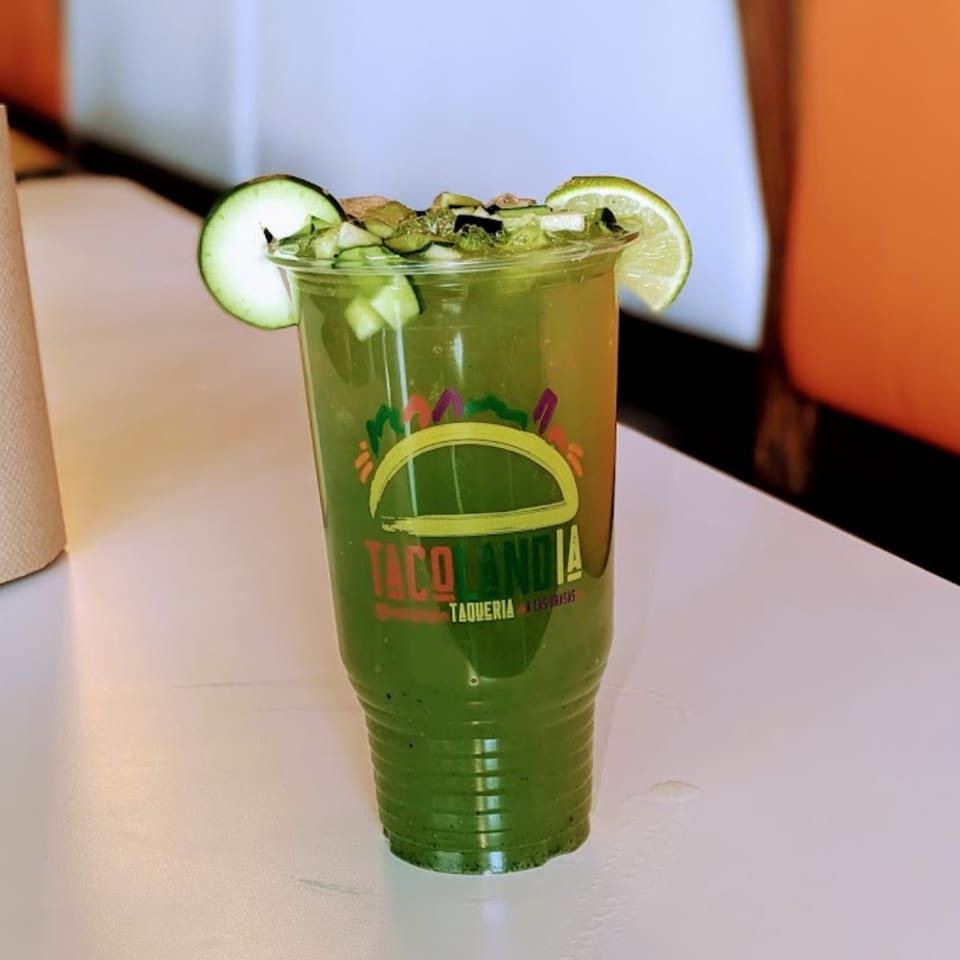 Tacolandia's green drink with cucumber slices and lime on a white table.
