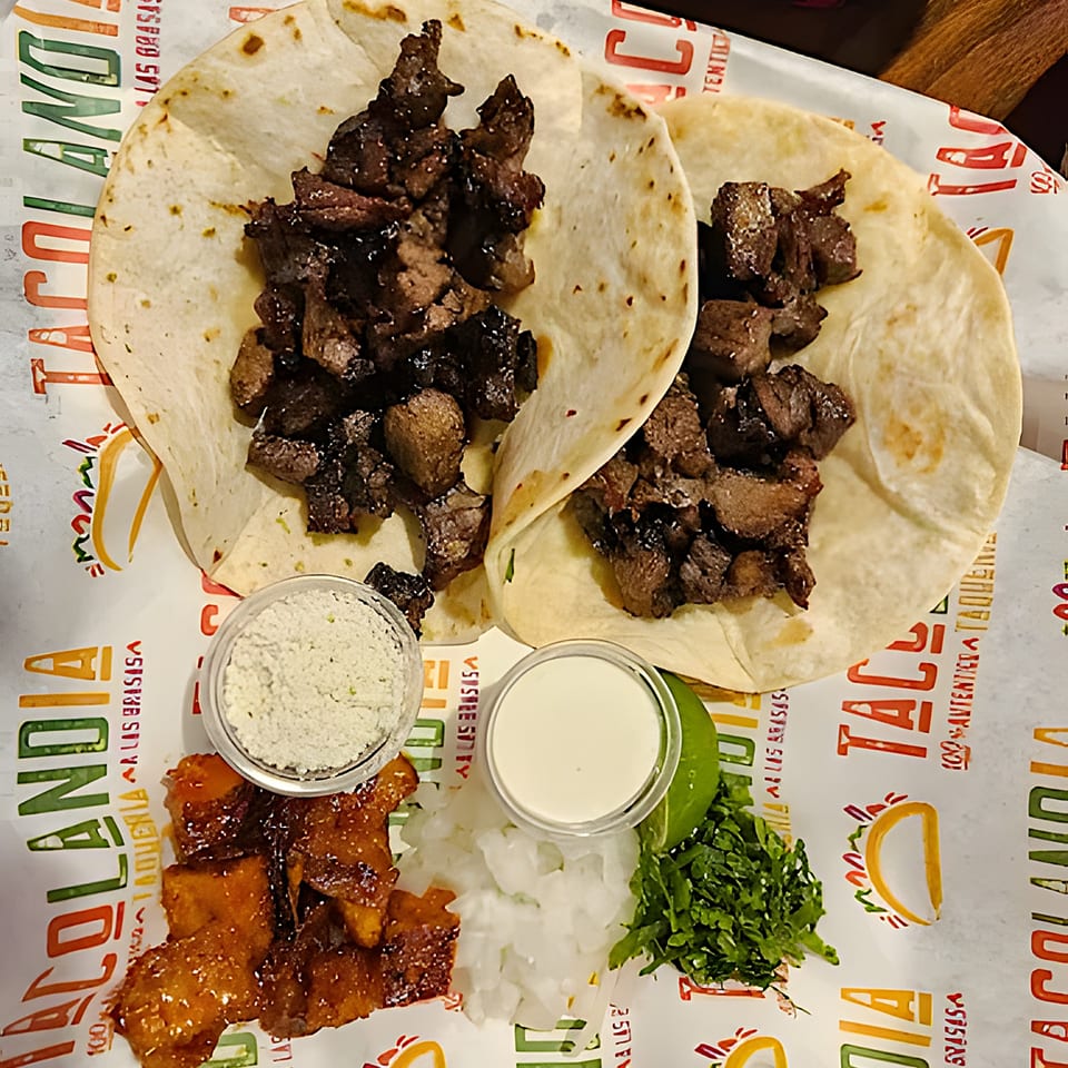 Two Tacolandia tacos with meat, sides of cilantro, onions, lime, and sauces on branded paper.