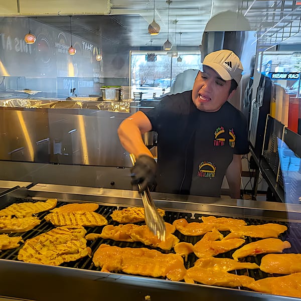 A Tacolandia chef in a cap and black shirt grilling chicken behind a glass counter.