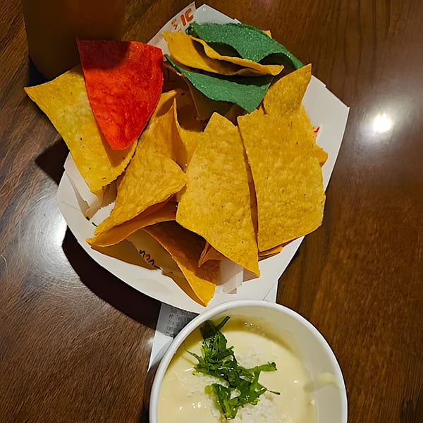 Colorful tortilla chips in a white container with a side of queso dip on a wooden table.